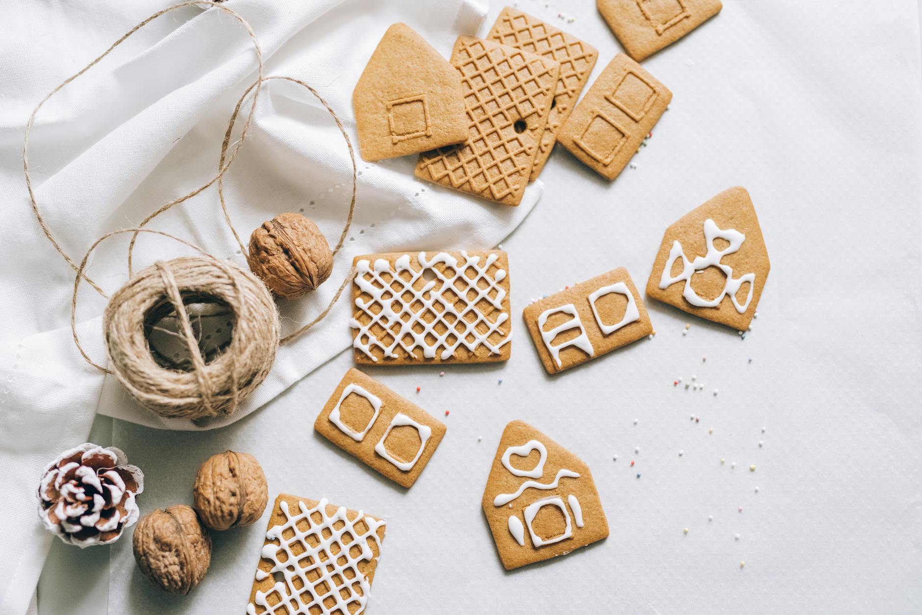 Festive flat lay with gingerbread house parts, walnuts, and string on a white cloth.
